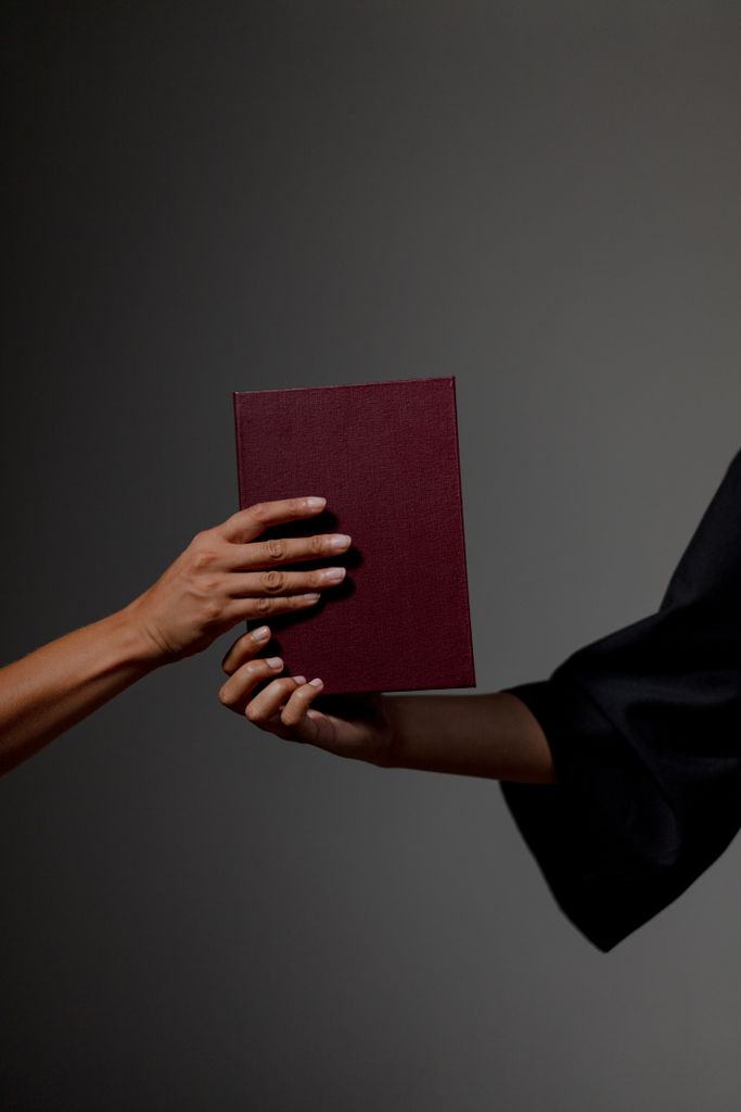 Close-up of two hands exchanging a diploma against a gray background, symbolizing graduation.